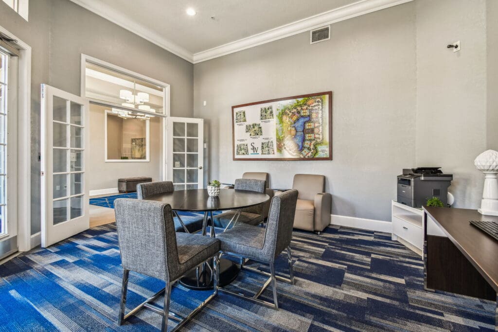 A modern resident workspace with a round black table surrounded by four gray upholstered chairs on a blue patterned carpet. Two beige armchairs sit against a light gray wall, above which hangs a large framed site map of the apartment community. To the right is a small desk with a printer and a white storage unit. Double French doors open to a bright adjoining room with contemporary lighting and additional seating, while natural light streams in through glass doors on the left, creating a clean and inviting atmosphere.